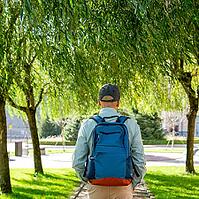 Man carrying backpack under willow tree in municipal park. Travelers explore nature and urban landscape. Concept of travel, adventure and leisure in summer sunshine. Rear view [IBR125492383]