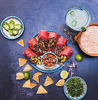 Top view of delicious burritos, pico de gallo, and more traditional food on dark grunge surface. Tortilla chips, sliced peppers, sauce and more on plates and clay pots. Authentic Mexican cuisine [IBR125492388]