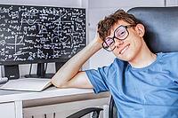 Cute teenage student in glasses sitting at a computer studying online, looking at the camera and smiling contentedly. [IBR125492406]