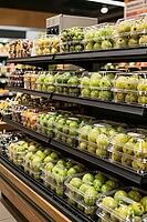 Fresh green fruits in transparent plastic clamshell boxes line dark supermarket shelves, showing modern food preservation and retail distribution, synthetic polymers encase organic agricultural goods in orderly multi level displays, highlighting the tensi [IBR125502126]