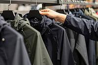 Technical outdoor apparel shopping defines functional garment curation, a hand inspects waterproof hooded jackets on rails inside an urban store, synthetic weather resistant fabrics define utilitarian trends in consumer culture, dark windbreakers line up  [IBR125502131]
