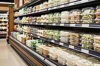 Industrial food preservation in modern commercial supermarkets, transparent plastic deli containers on chilled display shelves provide ready meals, refrigerated compartments hold sliced fresh produce alongside dairy delicacies and portioned snacks, linear [IBR125502137]