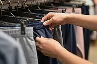 Tactile evaluation of activewear trousers on retail clothing racks highlights consumer demand for casual apparel and sensory product examination, a customer inspects the elastic waistband and knitted texture of dark blue fleece sweatpants displayed beside [IBR125502143]
