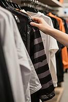 Tactile garment selection on retail clothing display stands inside a boutique, anonymous hand inspecting striped synthetic sportswear outerwear, physical merchandise evaluation contrasting fast digital commerce, monochrome windbreakers and casual shirts s [IBR125502145]