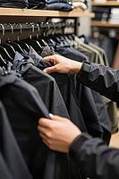Tactile evaluation of weatherproof synthetic apparel on garment racks in a retail store, hands inspect dark functional rain jackets for outdoor protection, the deliberate touch of waterproof textile membranes reflects modern consumer choices within urban  [IBR125502154]