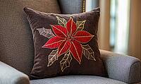 Dark chocolate pillow with bright red poinsettia and gold stitching, on a warm gray armchair, AI generated [IBR125502208]