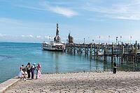 A small group gathers on the shore opposite an excursion boat and a pier reaching far into the sea, harbor with the Imperia statue, Constance on Lake Constance, Baden-Württemberg, Germany [IBR125521091]