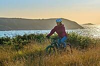 Active senior woman cycling with her electric mountain bike along the wild coast of Finisterre in southern Brittany next to Camaret-sur-Mer, France [IBR125521116]