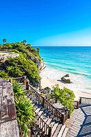 Vertical image of Playa Pescadores in Tulum, Quintana Roo, Mexico, showing wooden stairs and viewing platforms descending a rocky cliff covered in lush tropical vegetation to a sandy beach with turquoise Caribbean Sea waves under a clear blue sky [IBR125527063]