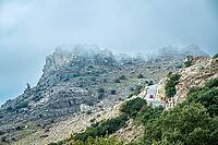 Rocky limestone mountain landscape with a winding paved road and small red car under misty, overcast sky in El Torcal near Antequera, Andalusia, Spain. Green shrubs and trees cover the slopes around the natural park roadway [IBR125527076]