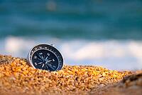 Conceptual photo, close-up of a compass lying in wet sand consisting of small crumbs of shells, against the backdrop of a blue calm sea [IBR125527077]