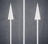 Two white arrows painted on a dark road surface, both straight ahead, separated from a vertical white line in the middle, representing directional traffic trails [IBR125536430]