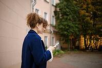 Woman using smartphone to order taxi on urban sidewalk beside apartment buildings and trees. Mobile communication, messaging and digital connectivity. Everyday city life and technology use [IBR125536432]