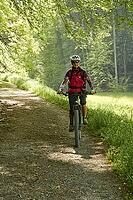 Mountain biker in red jacket rides through sunlit bright green spring forest. Frontal shot. Zurich Oberland, Switzerland [IBR125536456]