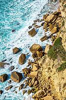 Rocky sea cliff coastline with large boulders, turquoise Atlantic water and breaking waves near Barbate in Andalusia, Spain. View from above shows textured sedimentary rock formations along the rugged shore in bright daylight [IBR125536461]
