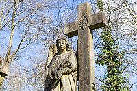 A weathered stone angel statue stands next to a large cross monument in the historic Highgate Cemetery in London under a blue sky [IBR125557550]