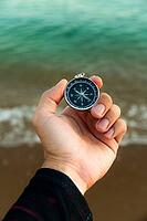 Pov of man hand holding navigational travel compass to find direction and next destination place. Concept of travel people and exploring. Beach and ocean in background [IBR125557610]