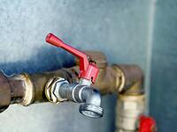 The red handle on a water pipe controls the water inlet. Gold and silver pipes horizontal and vertical in front of a gray wall [IBR125565996]
