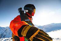 A guy in a ski suit holds skis in his hand and stands on a snowy mountain looking into the distance at the sun and a mountain range, ski resort and recreation development [IBR125565997]