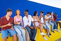 Diverse young adults sitting on a yellow wall, smiling and interacting with their smartphones against a vibrant blue background, symbolizing digital communication and social connection [IBR125566036]