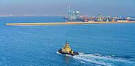 Manama, Bahrain - Jan 08 2024, Panoramic view of passing tugboat, a boat, in the background a commercial international port with cranes, pier, buildings and containers, in the daytime, Manama, Bahrain [IBR125566055]