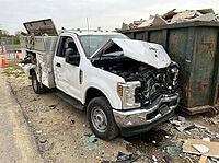A white utility pickup sits amid broken glass and rubble, its hood bent upward beside a metal container filled with demolition waste at a work site, AI generated [IBR125592668]