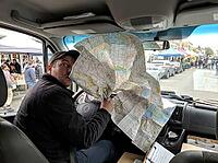 Inside the vehicle, a man grips a large folded road map across the dashboard as shoppers, market tents and parked cars line the street outside, AI generated [IBR125592684]