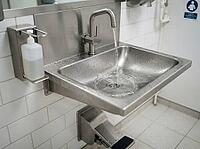 Water streams into the deep basin, while droplets bead on the metal surface beside a wall-mounted soap dispenser and white tiled walls, AI generated [IBR125592687]