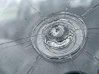 A close view of shattered automotive glass shows a central impact crater, concentric rings, fine spiderweb fractures, and scattered moisture, AI generated [IBR125592693]