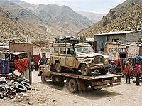 A dusty recovery truck hauls a weathered four-wheel-drive vehicle past adobe buildings, woven textiles and residents beneath rugged Andean peaks in a high-altitude rural settlement, AI generated [IBR125592705]