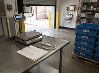 A stainless worktable holds a scale, probe thermometer, and receiving checklist beside reusable crates at a kitchen loading bay for food deliveries, AI generated [IBR125592715]