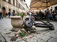 Broken masonry and scattered flowers cover the cobbled pavement as pedestrians pass cafe tables beneath broad umbrellas on a narrow street, AI generated [IBR125592717]