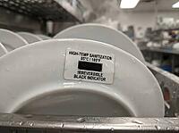 A water-speckled plate displays an irreversible heat indicator at 85degC, with dishware and stainless equipment visible in a commercial kitchen, AI generated [IBR125592724]