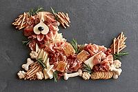 A playful dog-shaped charcuterie arrangement made from cured meats, cheese, breadsticks, crackers, nuts, and rosemary on a dark slate surface. AI generated [IBR125592737]