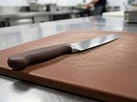 Close focus shows a brown prep board and polished blade on a stainless worktop, with kitchen equipment and a blurred worker nearby, AI generated [IBR125592748]