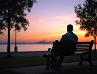 Person sitting on a bench, gazing at the sunset over calm waters, AI generated [IBR125621311]