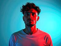 Studio portrait of a thoughtful young man with curly hair, illuminated by contrasting red and blue neon lights, creating a dramatic and colorful atmosphere, AI generated [IBR125621319]