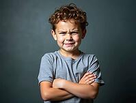 Young boy with curly hair crossing his arms and showing an angry expression on a gray background, AI generated [IBR125621323]
