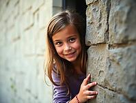 Portrait of a cheerful young girl hiding and smiling from behind a stone wall, AI generated [IBR125645914]