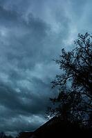 A bare tree and rooftop are silhouetted beneath heavy, dark blue-gray clouds [IBR125657365]
