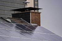 Solar panels cover a house roof beside a brick chimney and metal flue in daylight, with building walls visible in the background [IBR125657366]