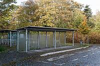 Metal shelters with glass panels stand beside a paved parking area, with fallen leaves on the ground and trees showing autumn colors in the background [IBR125657373]