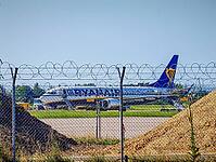 Airplane of the airline Ryanair, a white passenger aircraft with blue-yellow lettering and blue tail unit with yellow harp standing on the apron and being handling with vehicles and gangways, plus a wire mesh fence with barbed wire in the foreground, pile [IBR125657384]