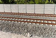 Railway tracks cross a gravel bed in front of a long sound barrier, with green treetops visible beyond [IBR125657403]