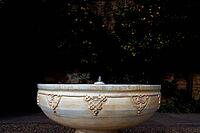 An ornate stone fountain bowl stands in the light against a dark garden surrounded by trees, Alhambra, Granada, Granada province, Andalusia, Spain [IBR125657409]