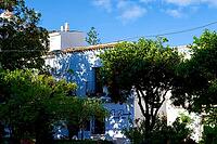 A blue and white house lies partially hidden behind lush trees, its leaves casting shadows in bright sunlight, Estepona, Costa del Sol, Andalusia, Province of Malaga, Spain [IBR125657431]