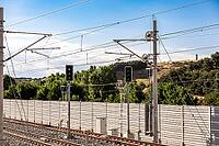 Railway signals and overhead electrical lines stand above tracks bordered by a sound wall and green hillside [IBR125657440]