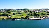 Bird's eye view of the Meersburg ferry dock on Lake Constance in a sunny summer day. A modern car ferry is just leaving the harbor basin, leaving a white spray trail in deep blue water. The background features lush green vineyard slopes, trees, and the ci [IBR125659392]