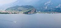 A wide view across Lake Garda toward a lakeside town and rugged green peninsula, with a motorboat crossing the water, Torbole, Lake Garda, Trentino, Italy [IBR125659411]
