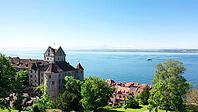 Picturesque view of the medieval Meersburg Castle on a sunny summer day. The historic fortress with its characteristic stepped gables and defence towers stands tall above the deep blue Lake Constance. A natural frame of green leaves in the foreground adds [IBR125659421]